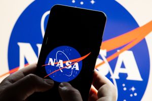A person holds a smartphone displaying the NASA logo in front of a larger NASA logo.
