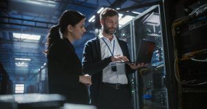 Two IT professionals review a laptop while standing in a data center server room.